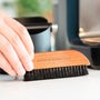 Afbeelding van een hand die een houten borstel met zwarte haren vasthoudt, met de tekst "BARISTA-ESSENTIALS" erop gegraveerd. De borstel wordt gebruikt om koffiedik van een werkblad te vegen.